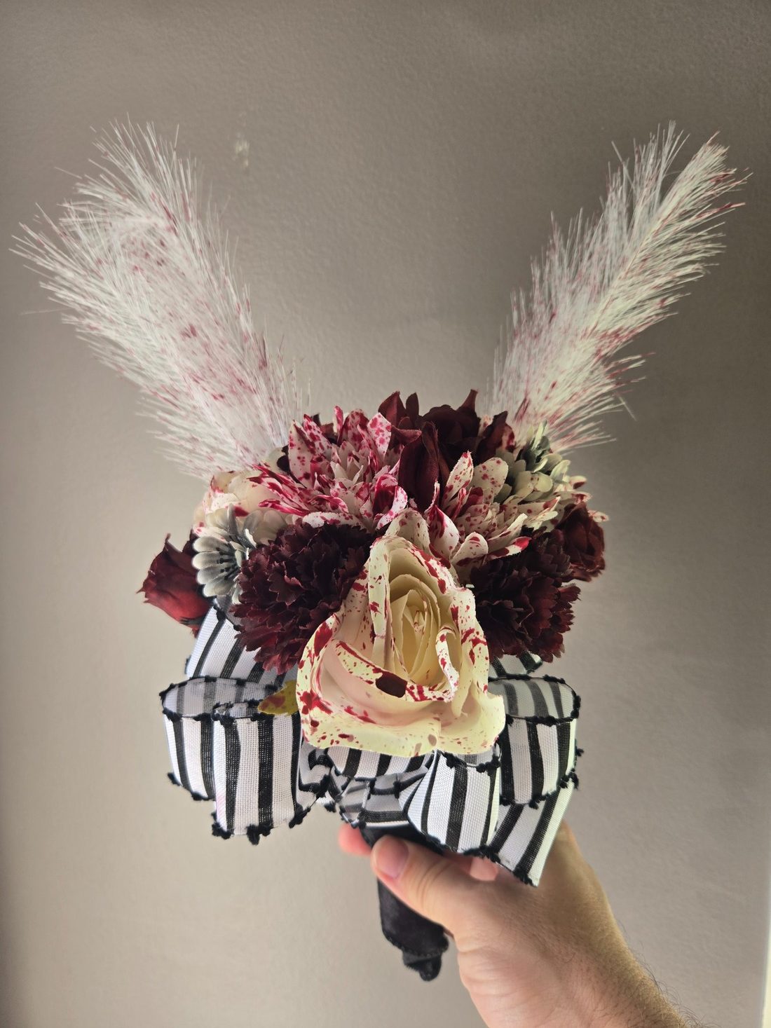 Our Bloody Bouquet is a hauntingly pretty mix of blood-splattered cream roses that GLOW IN THE DARK, deep burgundy carnations, and speckled pampas plumes, tied up with a bold black-and-white striped bow. Perfect for spooky centerpieces!